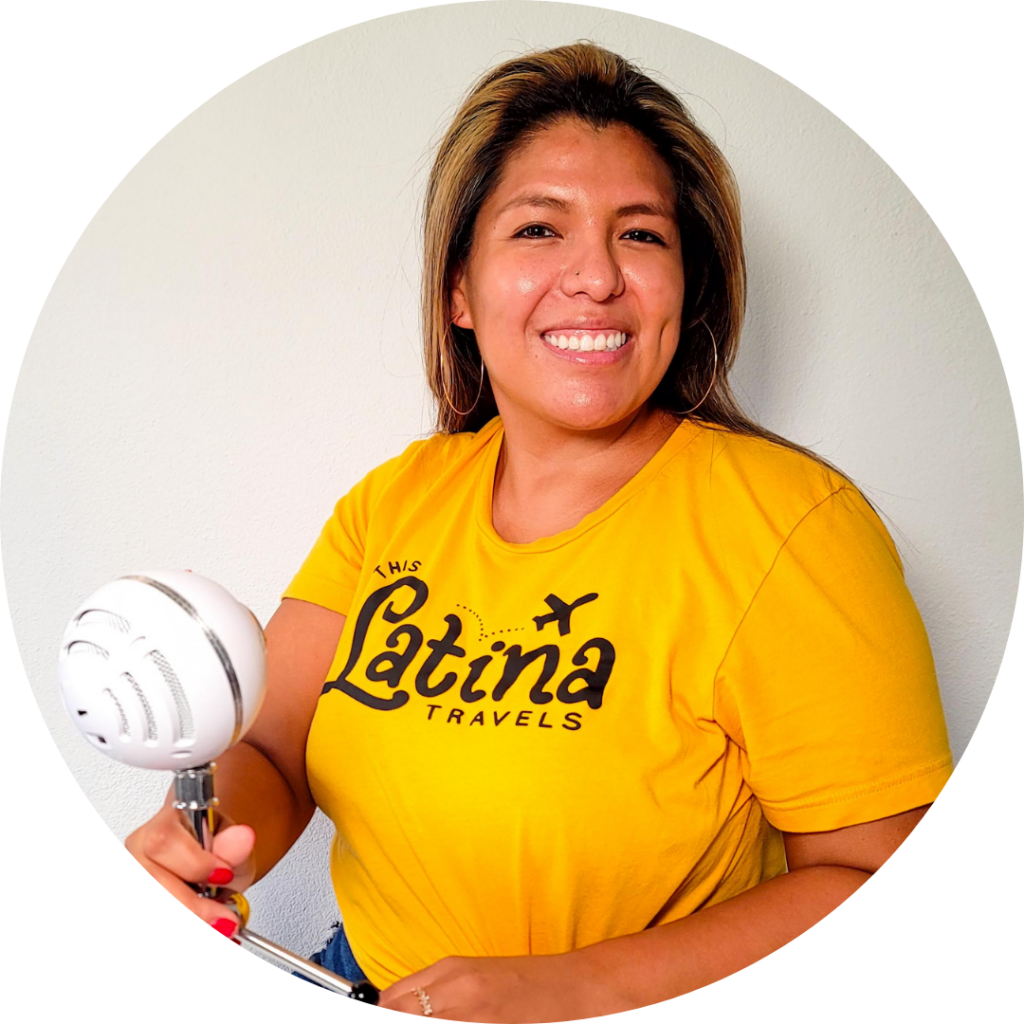 Bianca Alba, holding a white microphone and wearing her yellow This Latina Travels shirt, smiles at the camera