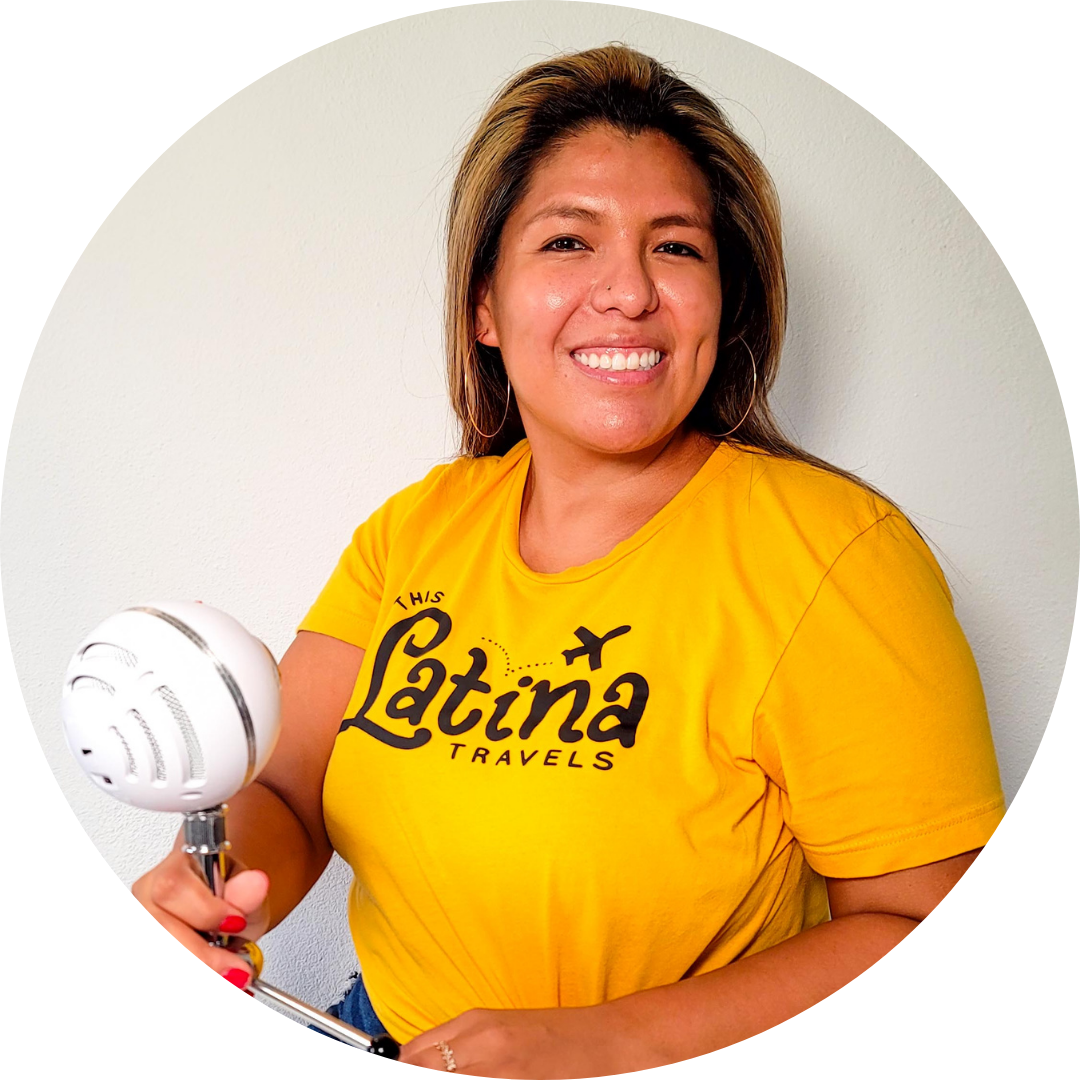 Bianca Alba, holding a white microphone and wearing her yellow This Latina Travels shirt, smiles at the camera
