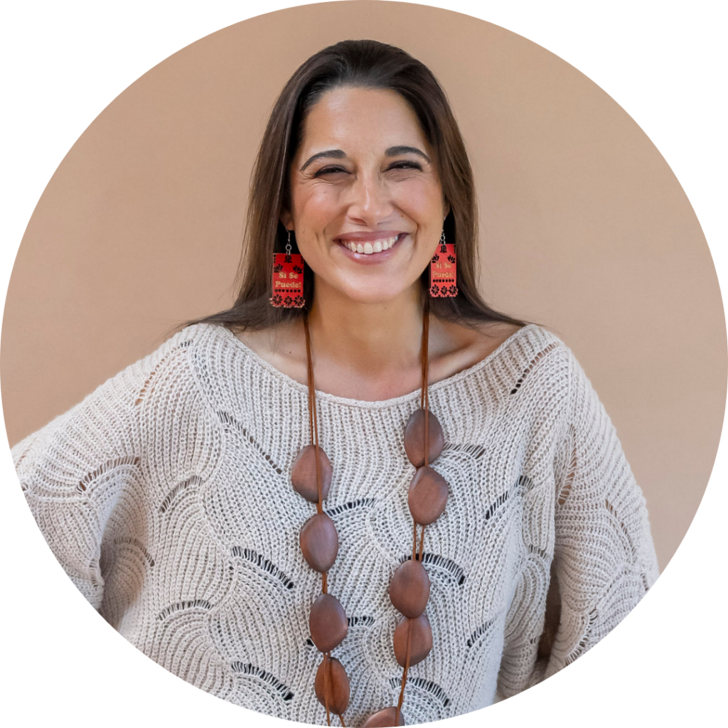 Mónika Aldarondo smiles at the camer in a light beige sweater with dangling earrings and long wooden necklace