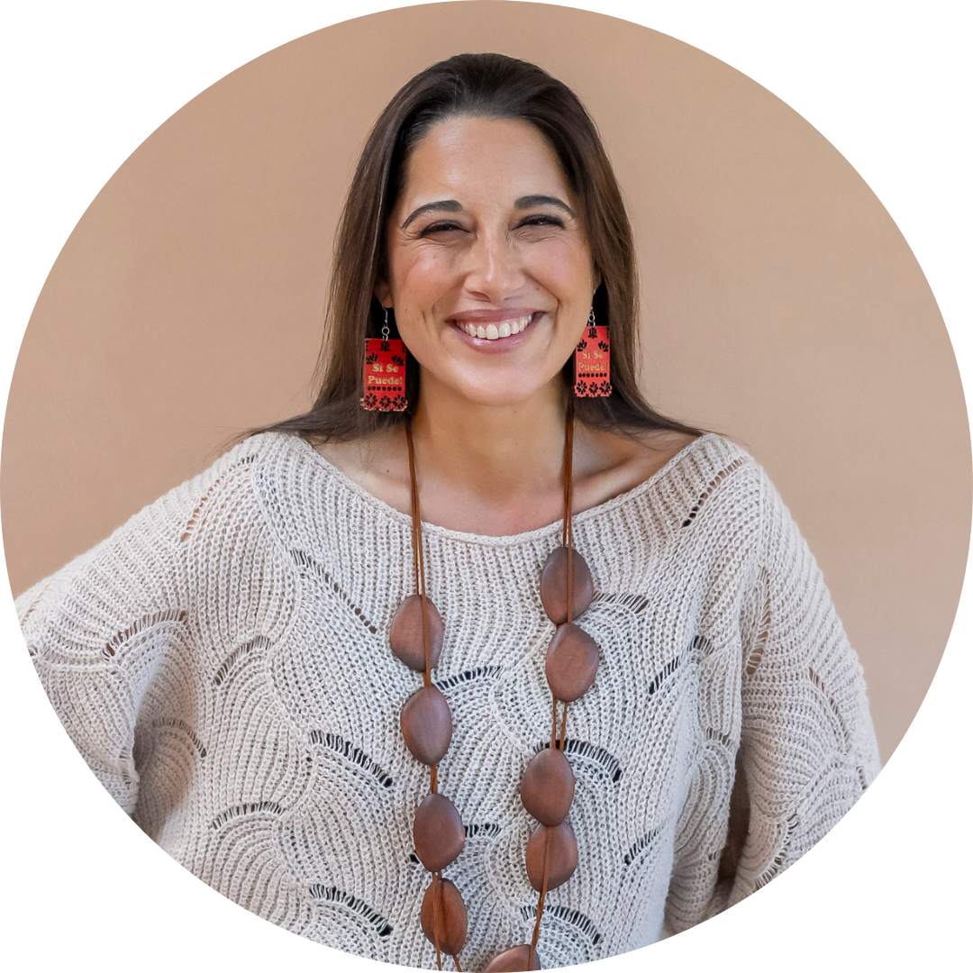 Mónika Aldarondo smiles at the camer in a light beige sweater with dangling earrings and long wooden necklace
