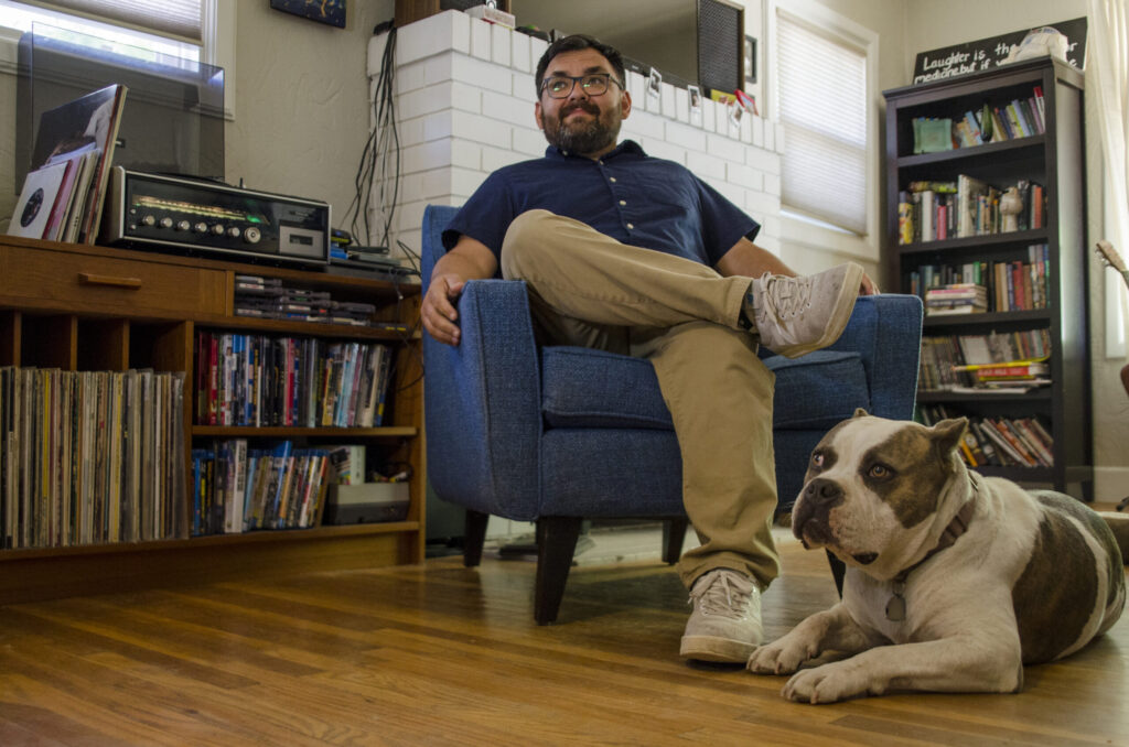 Johnny Flores sitting in a blue chair in his living room next to a vintage record player and records, his trusty pit bull down at his feet.
