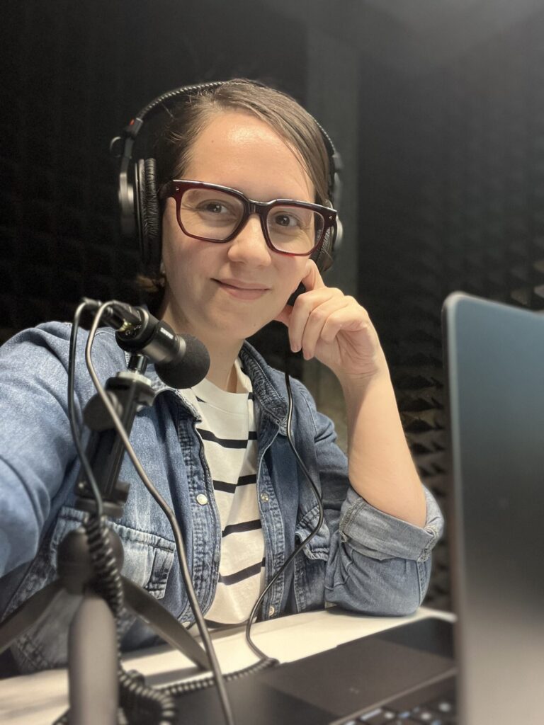 The author Analía Gomez Vidal wearing glasses and a headset, sitting casually at a microphone and computer looking ready to record a podcast episode.