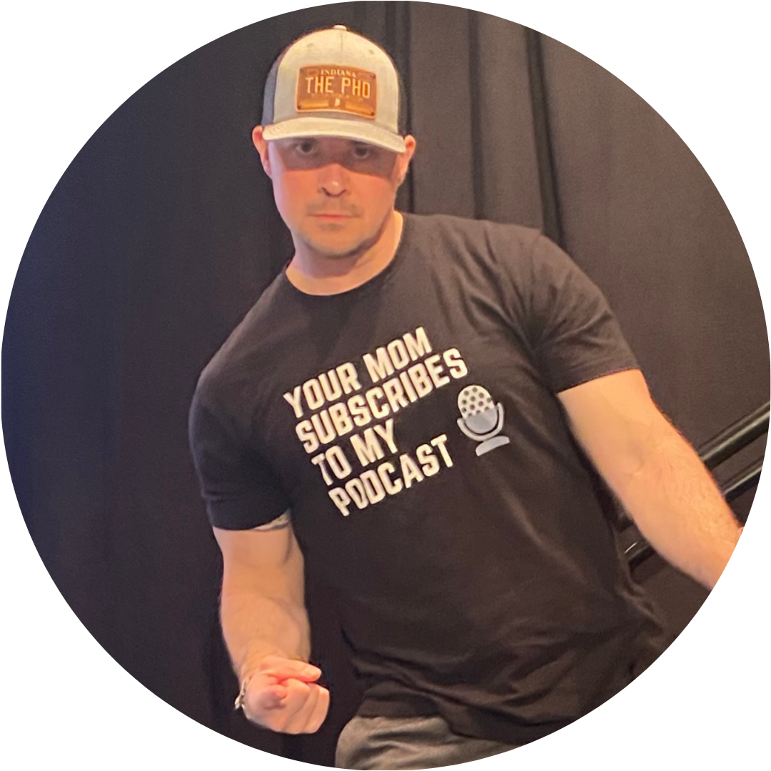 Adam Schauble leans away from a lectern pointing at the camera. He wears a light beige baseball cap that say PhD and a black t-shirt with block lettering that reads Your Mom Subscribes to My Podcast