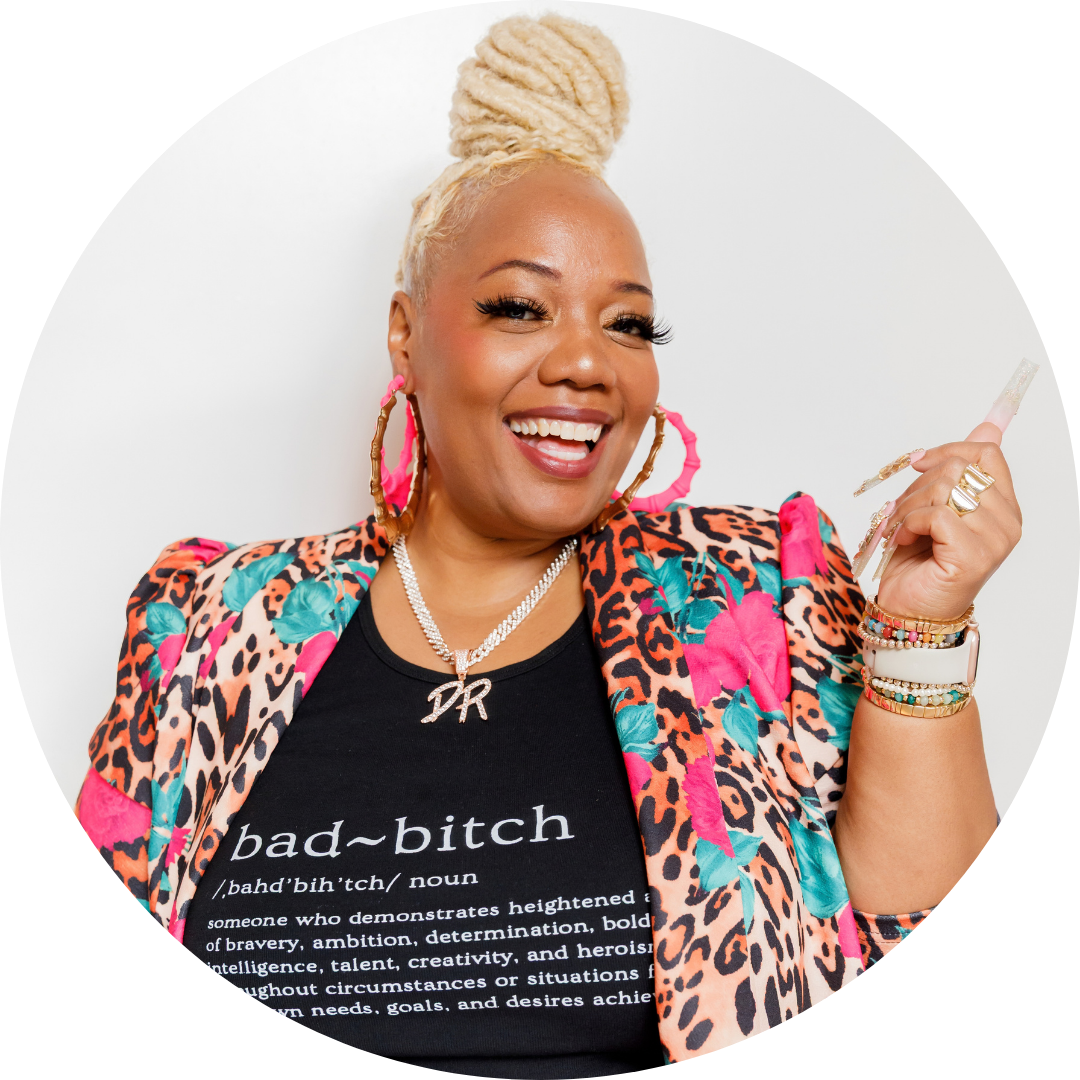 Dr Syreeta Rios Smiles at the camera wearing a pair of large pink hoop earrings, a colorful blazer, and a black shirt underneath with the definition of "bad bitch"