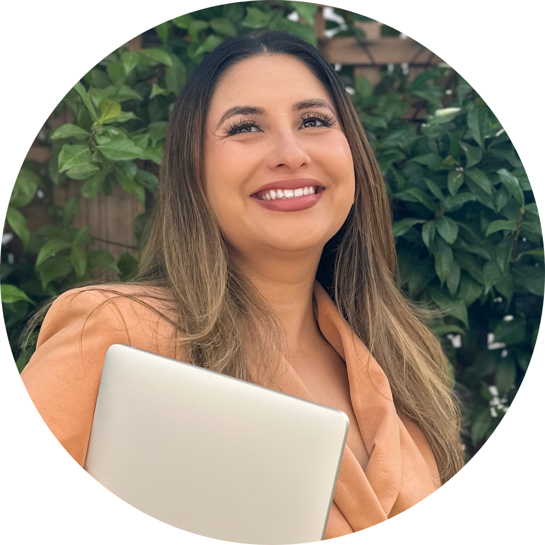 Jessica Lynn Rojas looks up above the camera, smiling while standing in front of some greenery and holding her laptop