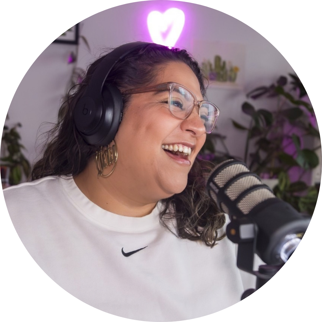 Kita Zuleta wearing glasses and a white sweatshirt laughing into a microphone while a pink neon heart glows above her head